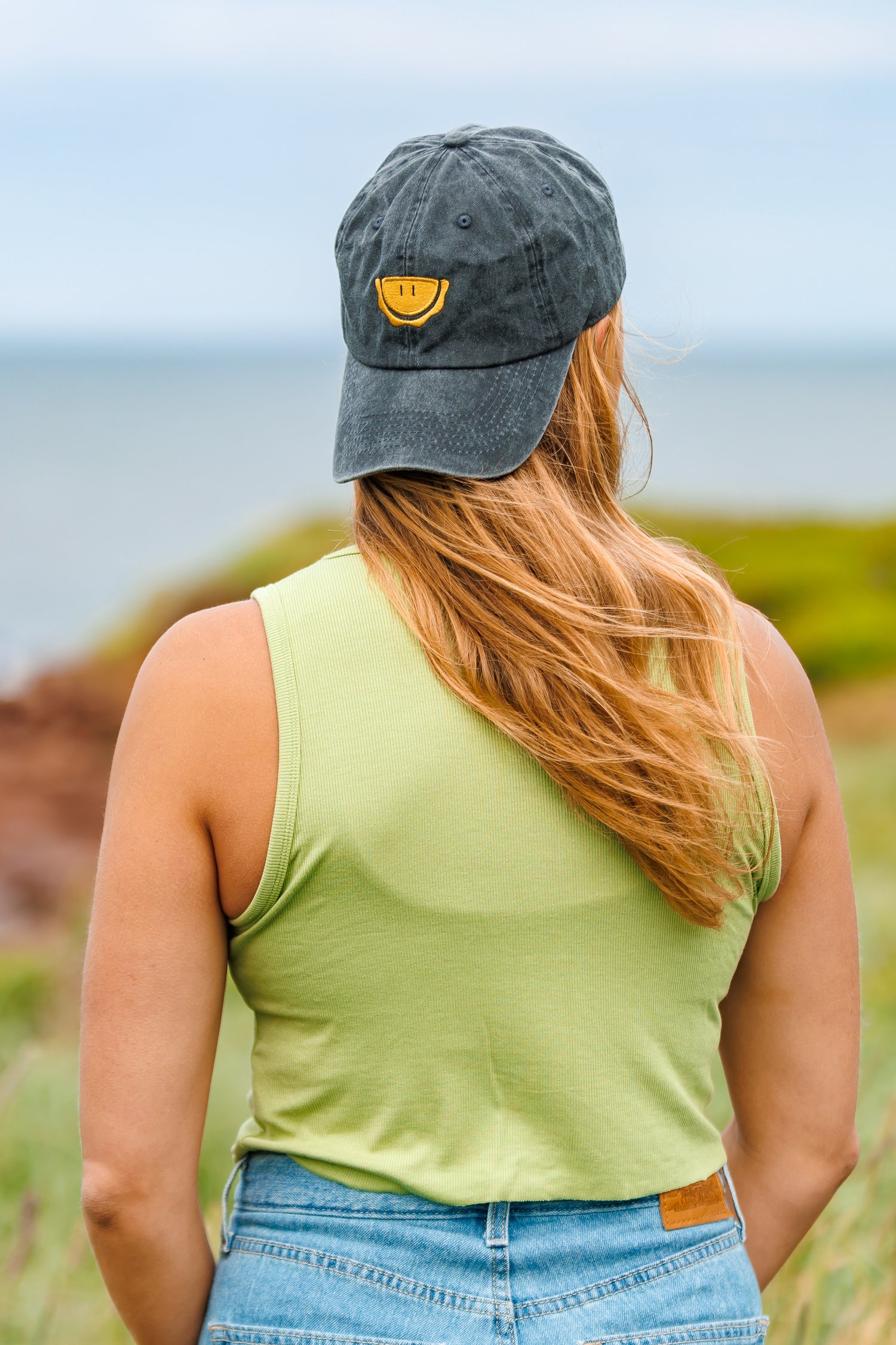 Blonde woman in a green shirt and jeans wears her The Handpie Co. merch hat backwards standing near red cliffs in PEI